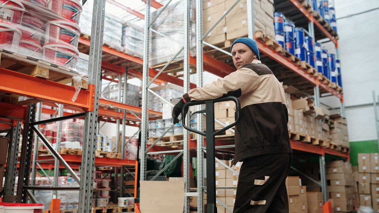 Warehouse worker operating a pallet jack among stacked boxes and metal shelves in an industrial setting.